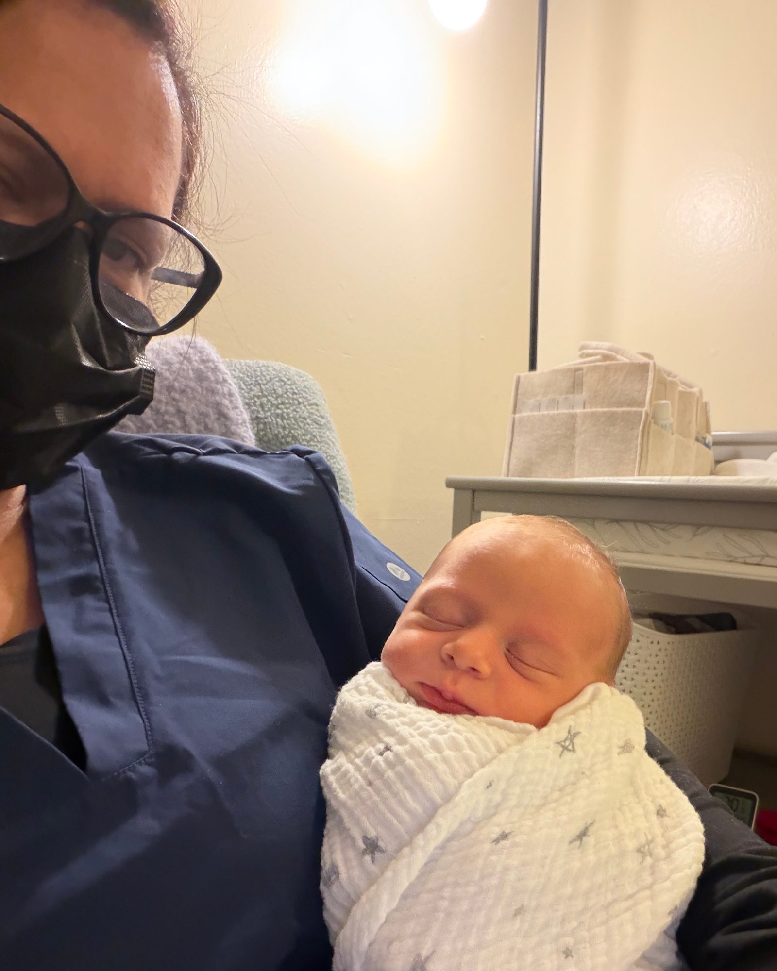 Dorah in navy scrubs and a face mask, holding a sleeping swaddled newborn